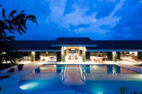 A luxurious poolside at twilight with a large open building, surrounded by seating areas and lush greenery, under a dramatic evening sky.