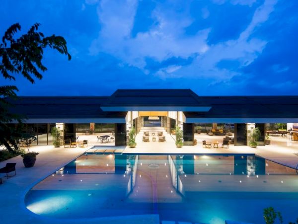 A luxurious poolside at twilight with a large open building, surrounded by seating areas and lush greenery, under a dramatic evening sky.