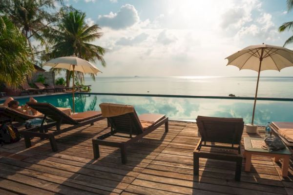 A serene tropical scene with lounge chairs, umbrellas, and palms by a pool overlooking the ocean under a bright sky.