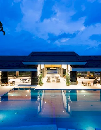 A luxurious building with large glass windows is illuminated at night, overlooking an expansive swimming pool and surrounding seating areas.