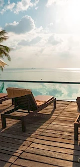 Poolside view with loungers, umbrellas, palm trees, and ocean in the background under a bright sky.