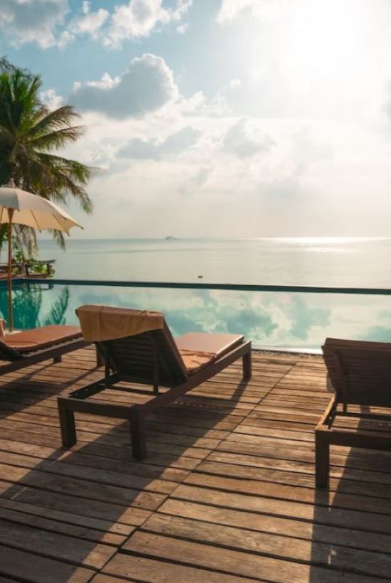A serene beachfront scene with lounge chairs, umbrellas, and a pool, overlooking the ocean under a bright, cloudy sky.