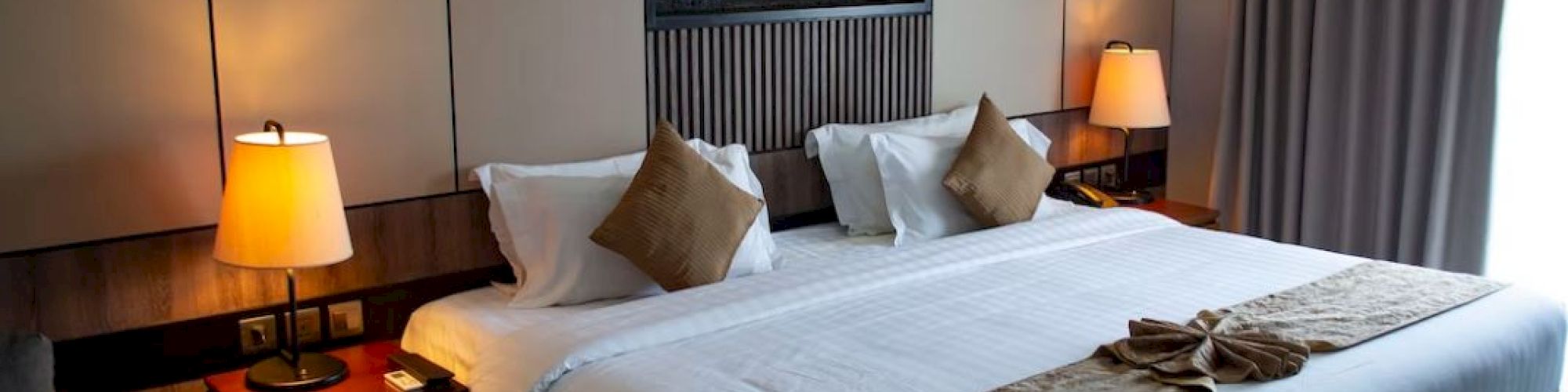 A neatly arranged hotel room with a large bed, decorative pillows, bedside lamps, and a window with closed curtains.