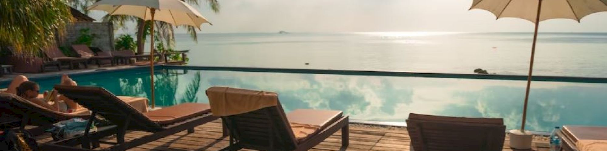 A serene poolside scene with lounge chairs, umbrellas, and palm trees overlooking a calm body of water and a bright sky.