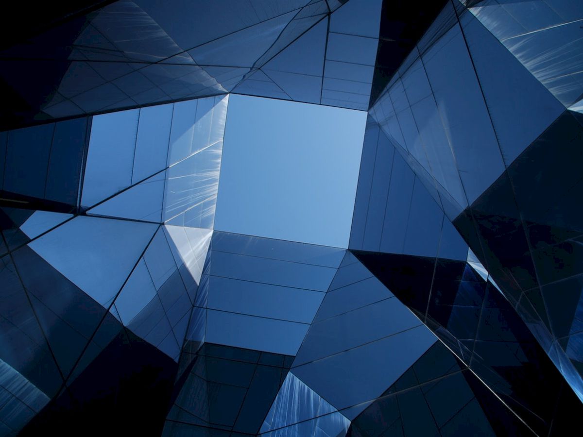 Abstract geometric structure with reflective glass panels forms a symmetrical pattern, viewed from below against a clear blue sky.