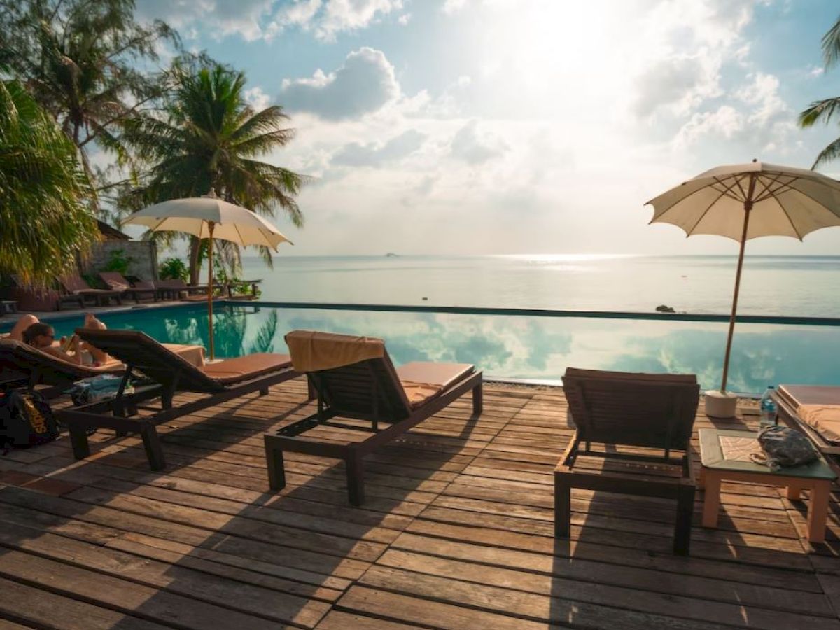 A serene poolside view with lounge chairs, umbrellas, palm trees, and a calm seascape under a partly cloudy sky.