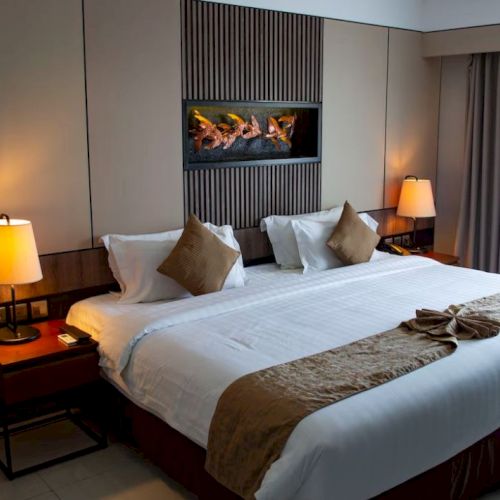 A cozy hotel room with a large bed, lit lamps on nightstands, decorative pillows, a wall art panel, and a window with curtains.