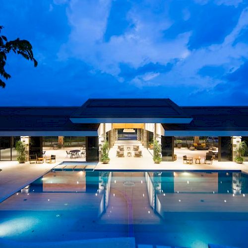 The image shows a modern building with a well-lit exterior and a large swimming pool in front, set against a twilight sky.