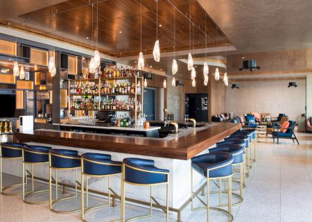 The image shows a modern bar with a wooden countertop, blue stools, hanging lights, and shelves stocked with bottles. There's seating in the background.
