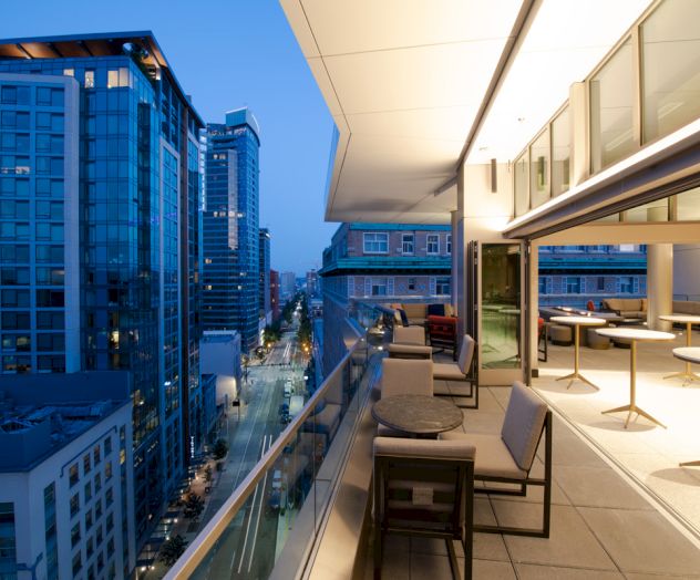 A modern cityscape featuring high-rise buildings and a well-lit, stylish balcony overlooking the street below at twilight.