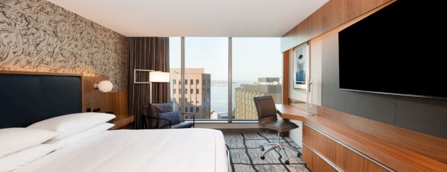 A modern hotel room with a large bed, desk, chair, and TV. There's a cityscape view through the window.