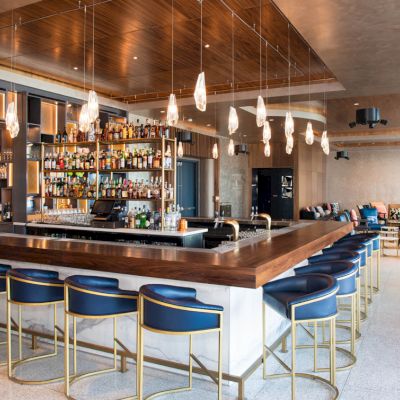 It's a stylish bar with blue stools, a wooden counter, hanging pendant lights, and a well-stocked shelf of bottles in a modern setting.