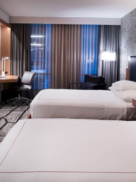 A modern hotel room with two beds, a TV, desk, and chair. The wallpaper is patterned, and the lighting is ambient and stylish.
