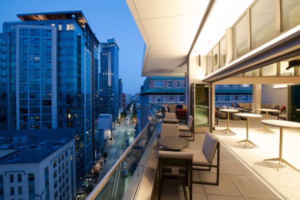 A modern terrace overlooking a cityscape at dusk, with tables, chairs, and buildings under a twilight sky.