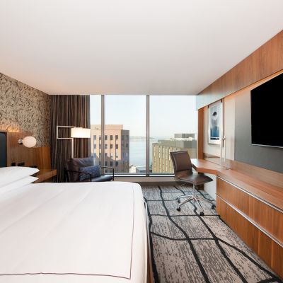 A modern hotel room with a large bed, wall-mounted TV, desk, and chair. A window offers a cityscape with buildings and a body of water visible.