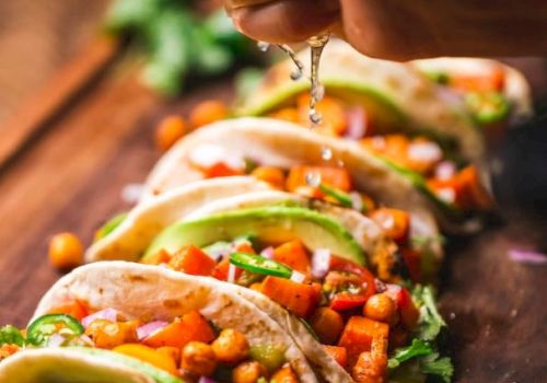 Five tacos filled with chickpeas, avocado, and vegetables are arranged on a wooden board while a hand squeezes lime over them.