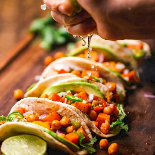 Five tacos filled with chickpeas, avocado, and vegetables are arranged on a wooden board while a hand squeezes lime over them.