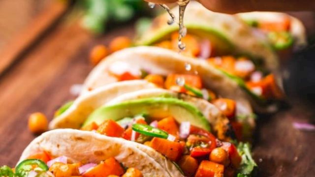 Five tacos filled with chickpeas, avocado, and vegetables are arranged on a wooden board while a hand squeezes lime over them.