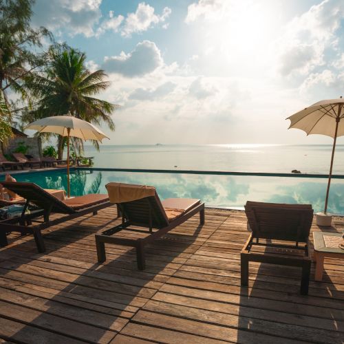 A picturesque tropical scene with lounge chairs, umbrellas, and a swimming pool overlooking a calm sea under a partly cloudy sky.