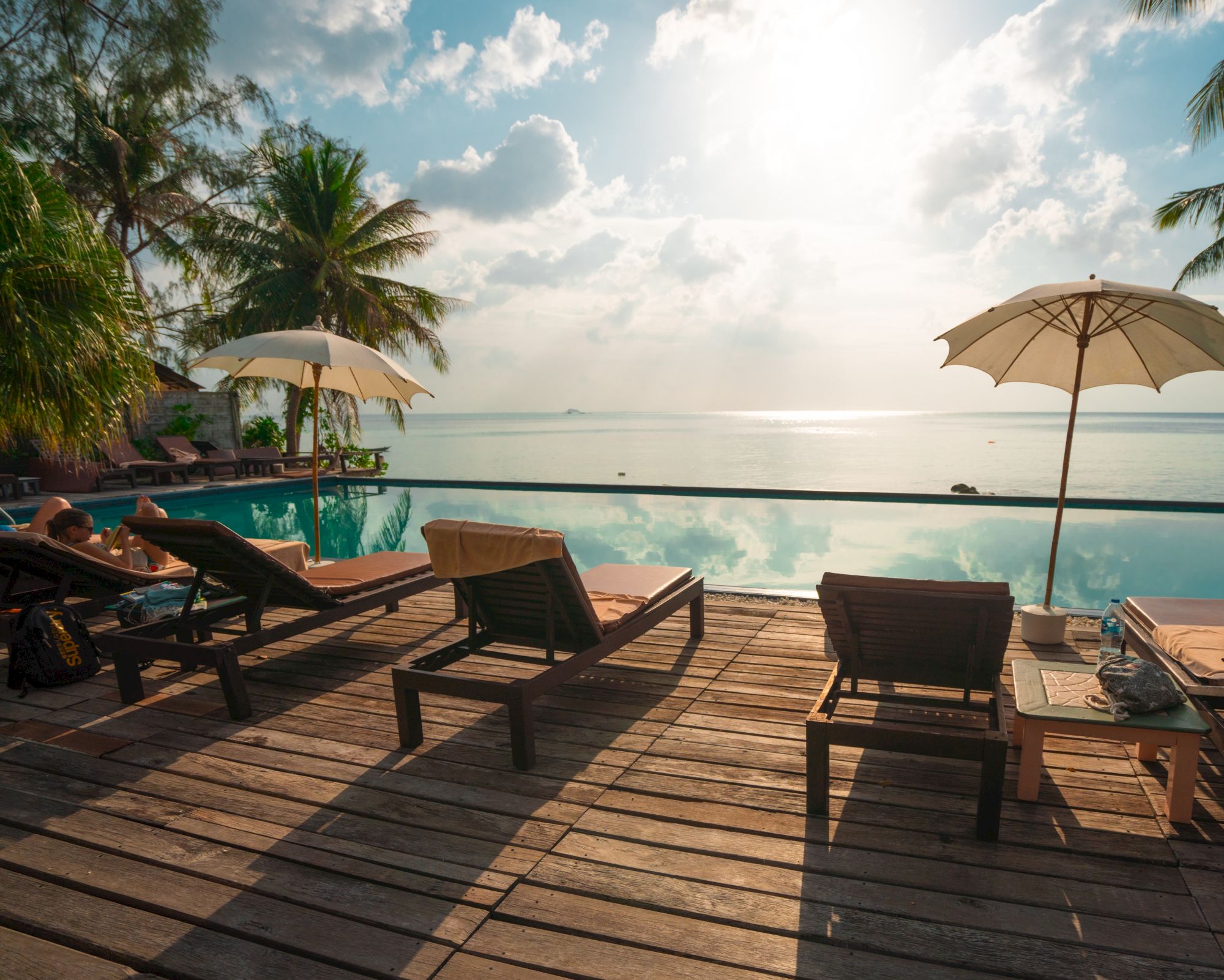 The image shows sun loungers and umbrellas on a wooden deck by an infinity pool, overlooking a serene ocean view under a partly cloudy sky.