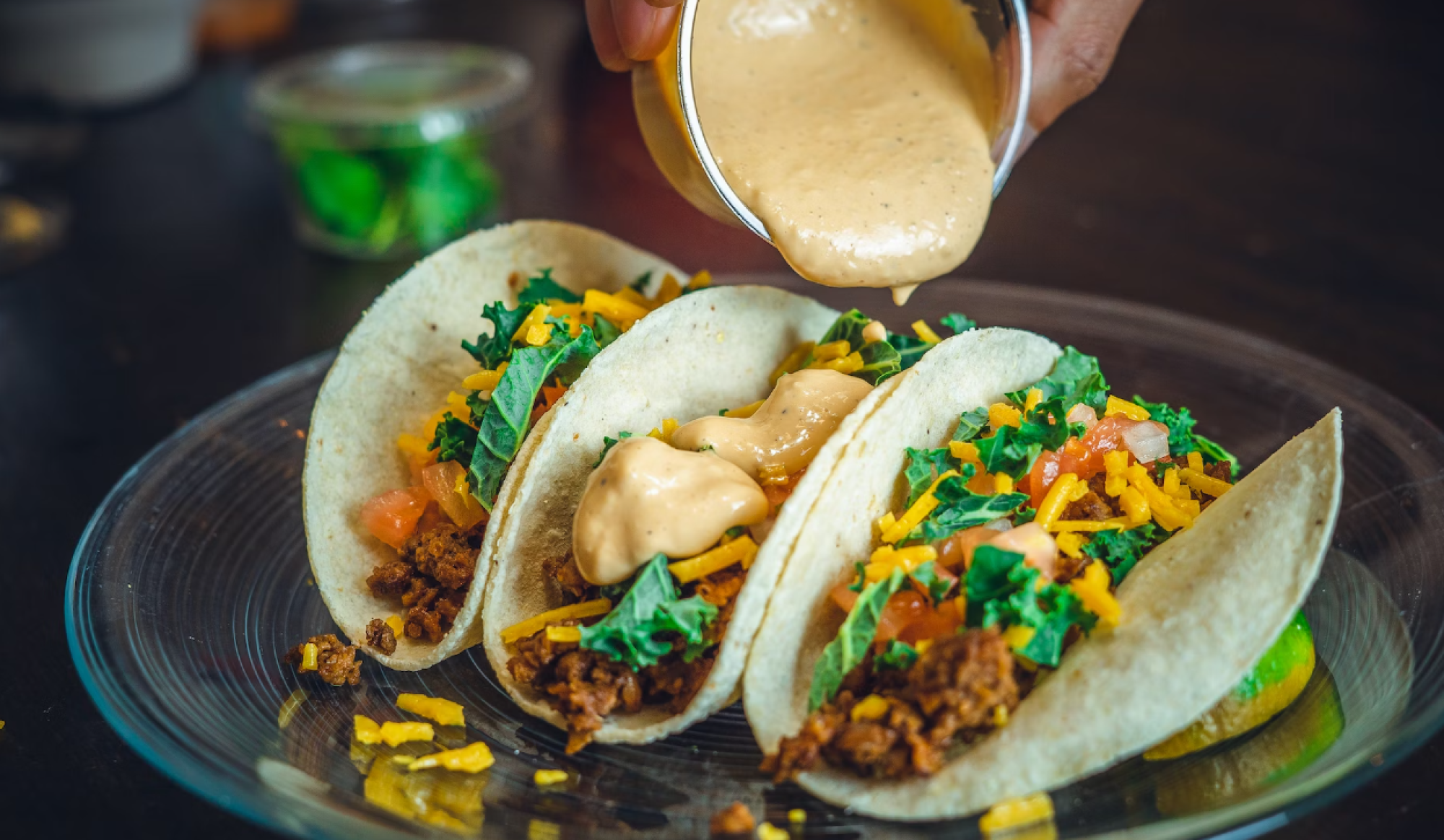 Three tacos filled with ground meat, lettuce, tomatoes, and cheese are topped with a creamy sauce being poured from a cup.