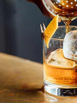A cocktail is being poured into a glass with ice, garnished with a twist of lemon peel, on a wooden surface.
