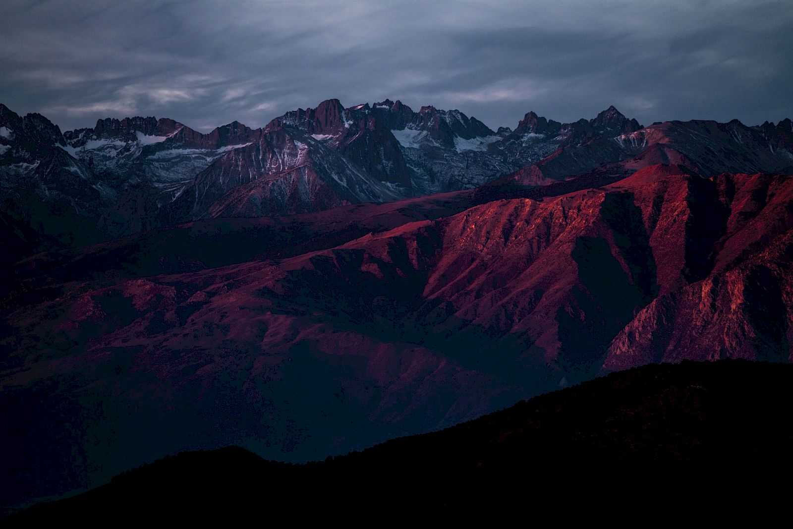 A mountain range with peaks dusted in snow, highlighted by dramatic red and purple lighting, under a cloudy sky.