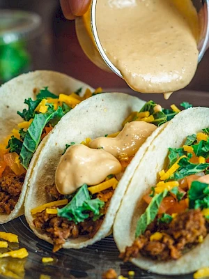 Three tacos filled with meat, cheese, and greens, being topped with a creamy sauce on a plate.