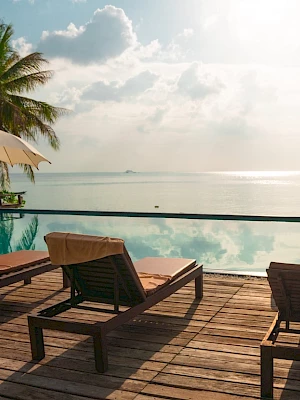 A serene poolside scene with lounge chairs and umbrellas overlooking a calm body of water, surrounded by palm trees under a partly cloudy sky.