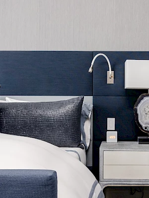 This image shows a modern bedroom with a blue headboard, pillows, a bedside table, a lamp, and a light gray wall.