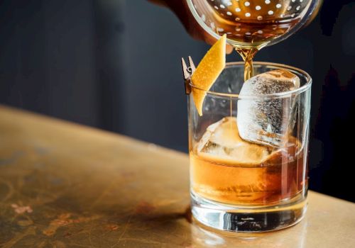 A drink being poured into a glass with ice and lemon peel garnish, on a bar counter.