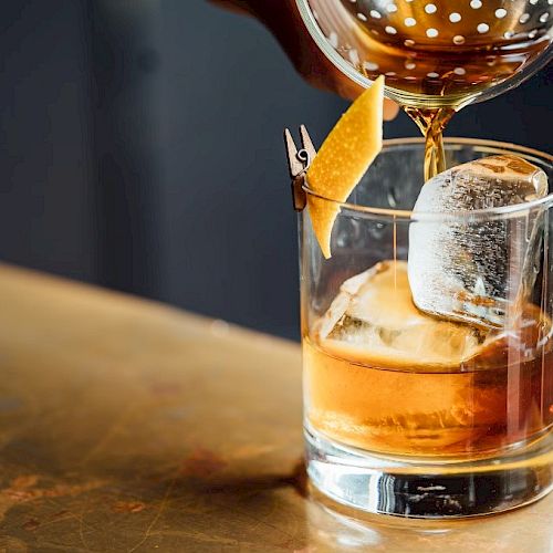 A drink being poured into a glass with ice and lemon peel garnish, on a bar counter.