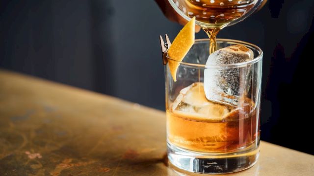 A drink being poured into a glass with ice and lemon peel garnish, on a bar counter.
