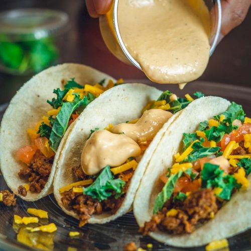Three tacos with ground meat, lettuce, tomatoes, cheese, and sauce being poured over them on a plate.