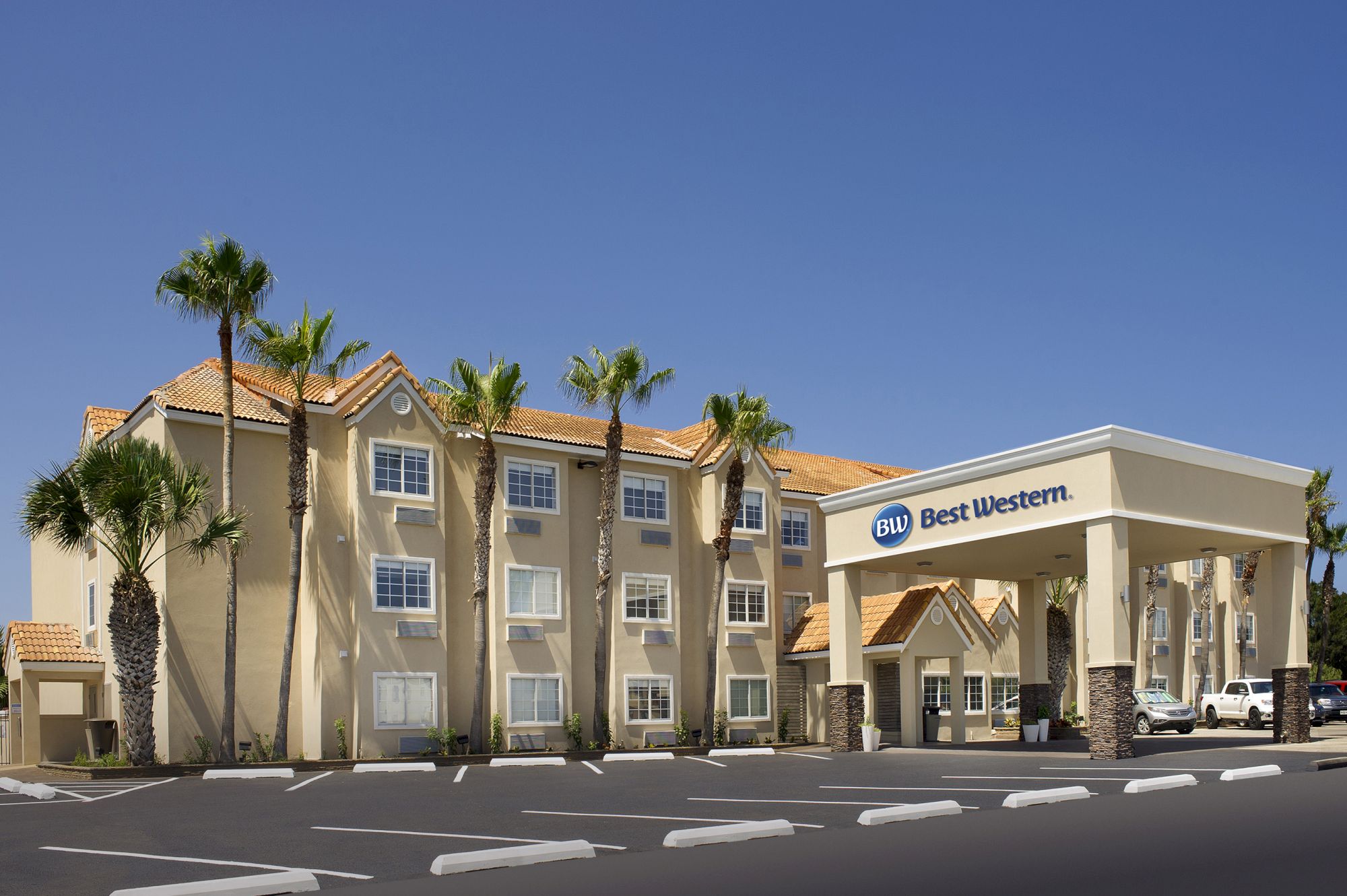 The image shows a Best Western hotel with palm trees, a clear sky backdrop, and a parking lot in the foreground.