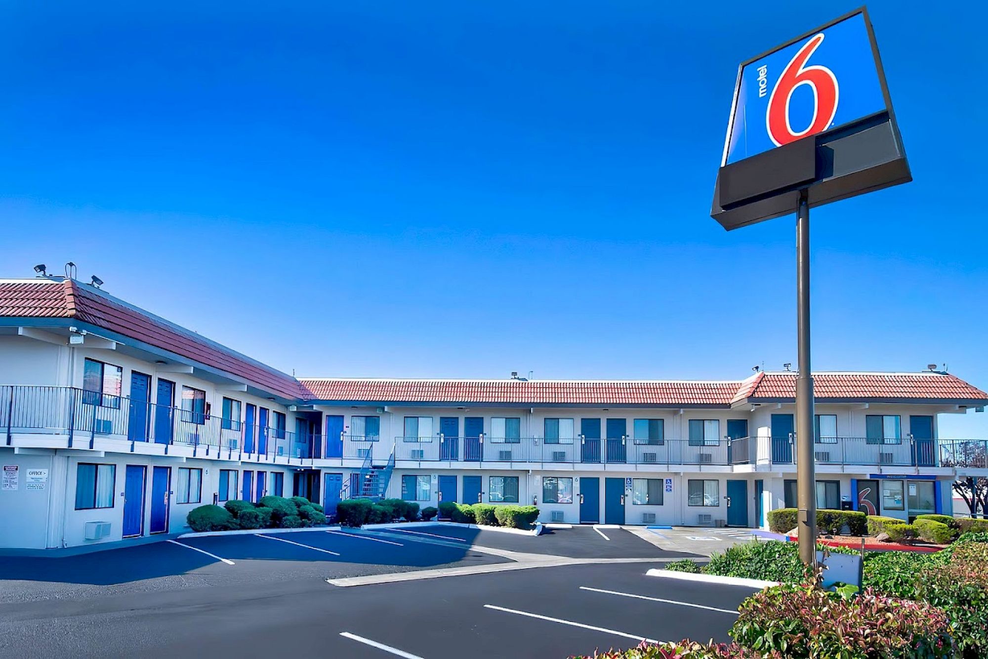 The image shows a motel building with a red and blue "6" sign, featuring a parking lot and clear blue skies.
