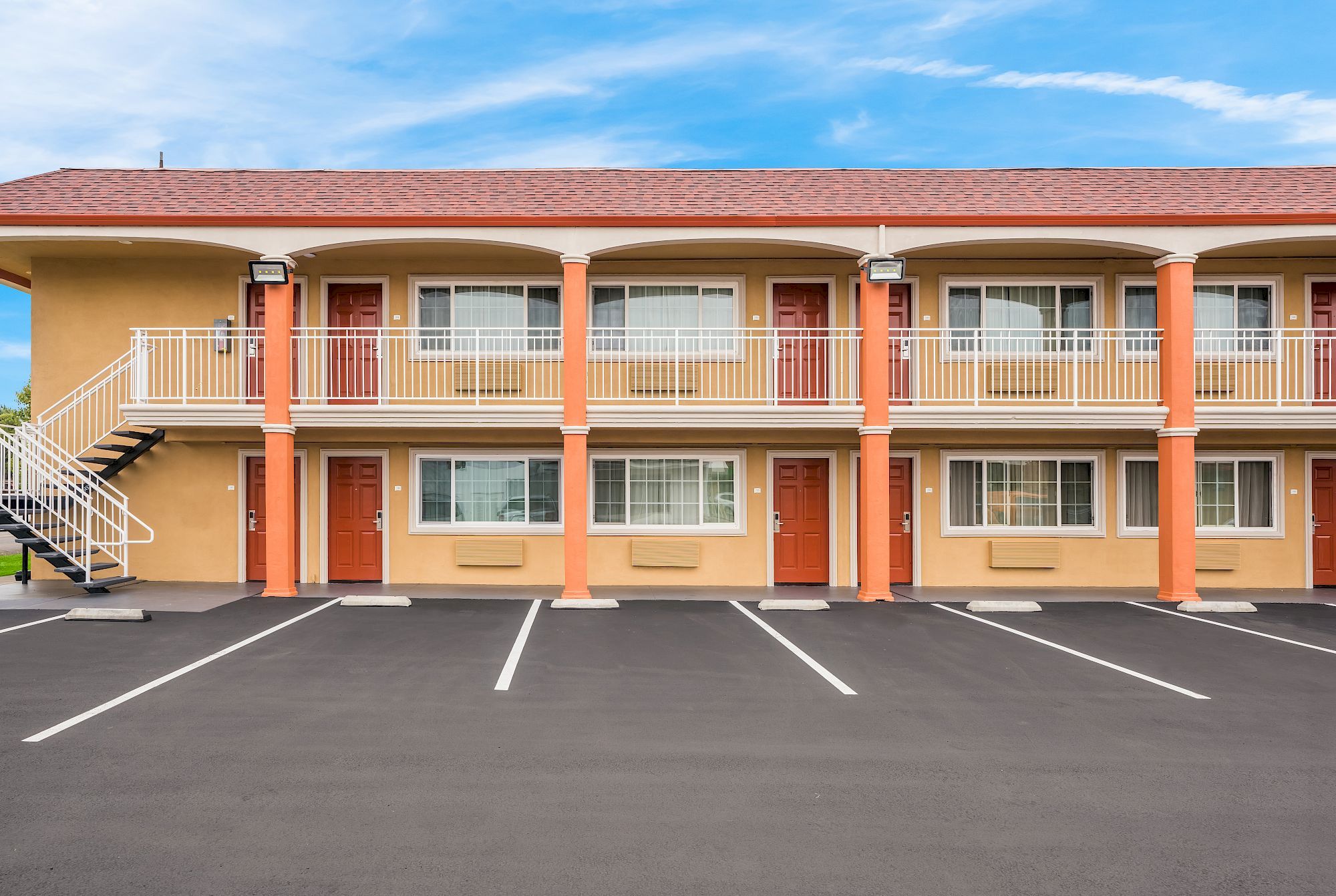 The image shows a two-story motel with beige walls, red doors, and empty parking spaces in front. The sky is blue with scattered clouds.