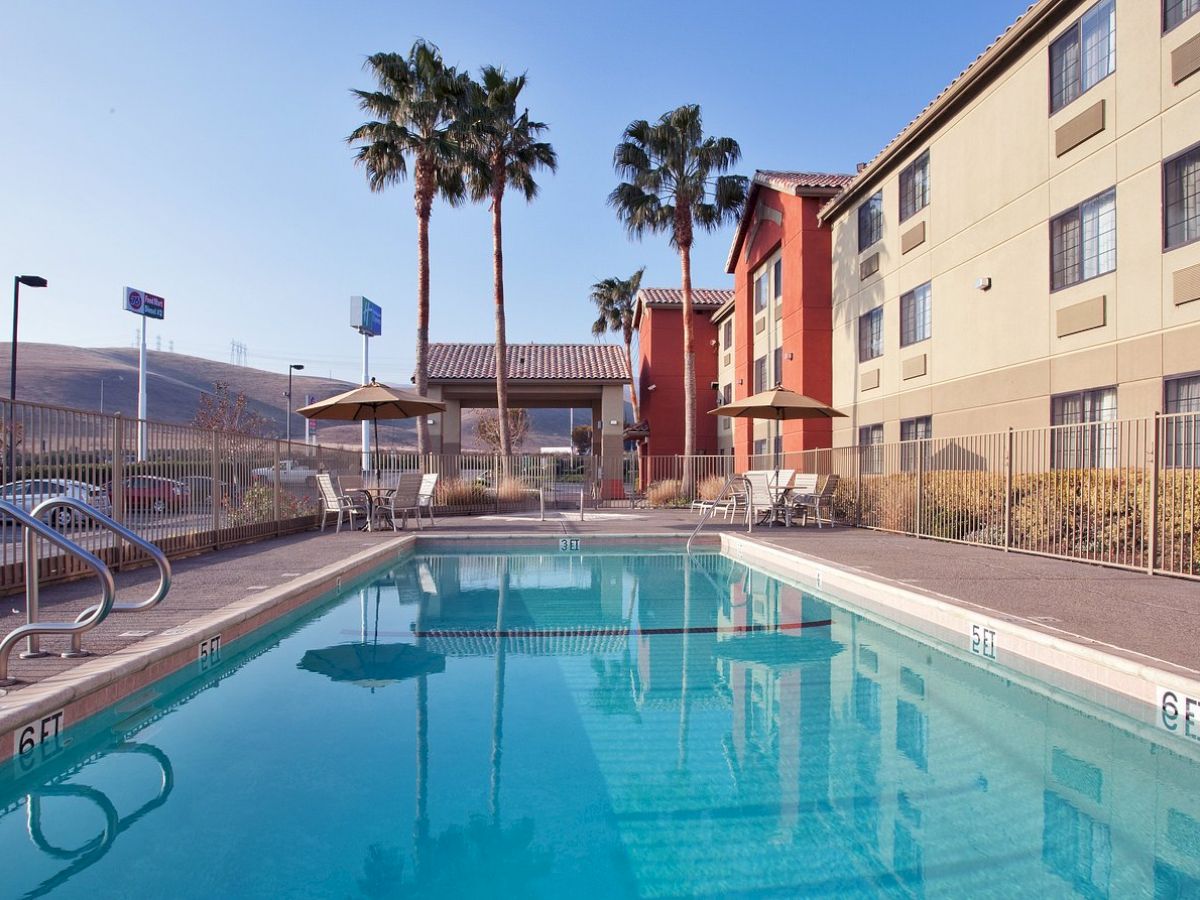 An outdoor pool surrounded by a fence, with palm trees and a multi-story building in the background, sunny day.