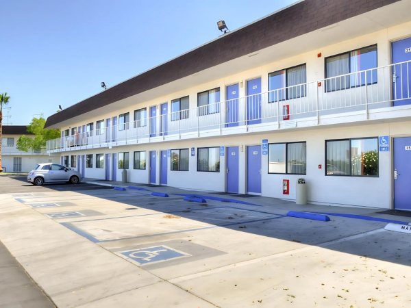 The image shows a two-story motel with exterior corridors, a row of doors, windows, parking spaces, and a car parked in front.
