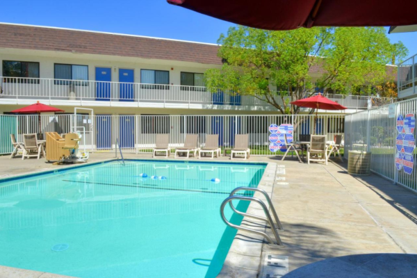 A motel pool area with lounge chairs, red umbrellas, and a two-story building in the background. The scene is sunny and inviting.