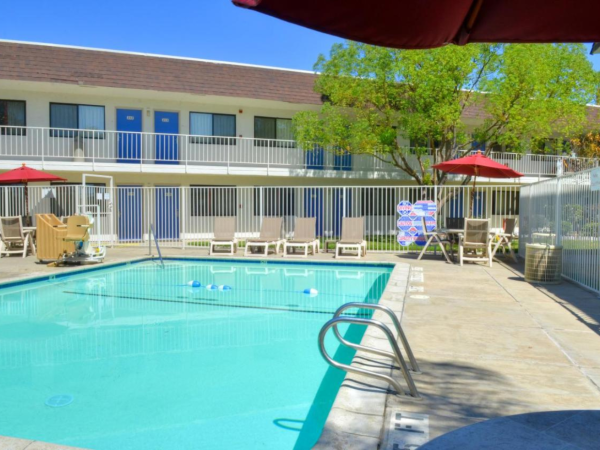 A motel pool area with lounge chairs, red umbrellas, and a two-story building in the background. The scene is sunny and inviting.
