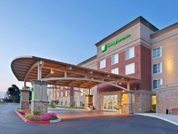 The image shows the exterior of a contemporary hotel building with a covered entrance and well-maintained landscaping.