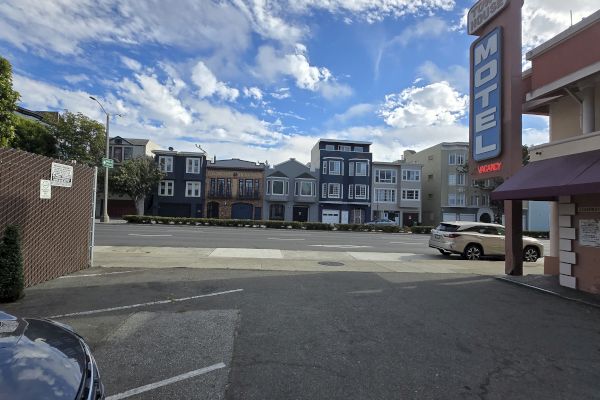 A parking lot with a motel sign on the right and colorful row houses across the street under a partly cloudy sky.