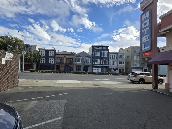 A parking lot with a motel sign on the right and colorful row houses across the street under a partly cloudy sky.