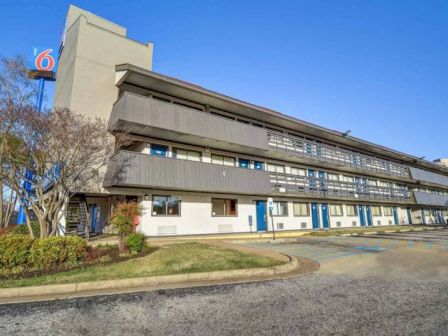 The image shows a three-story building with a Motel 6 logo, featuring outdoor corridors, accessible parking, and landscaped surroundings.