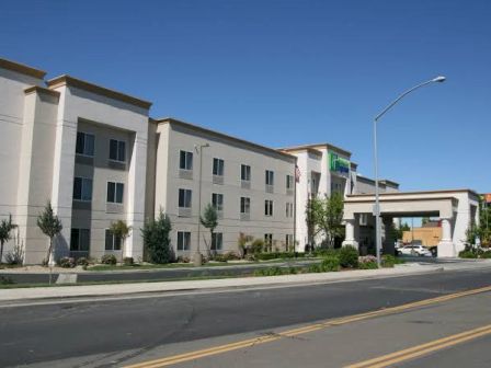 The image shows a modern, three-story hotel building with a driveway and a clear blue sky in the background.