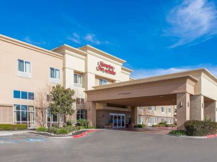 The image shows the exterior of a Hampton Inn & Suites hotel, featuring a welcoming entrance with a covered driveway and clear blue sky.