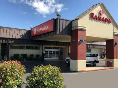 The image shows the entrance of a Ramada hotel, featuring a sign and a portico with a van parked in front.