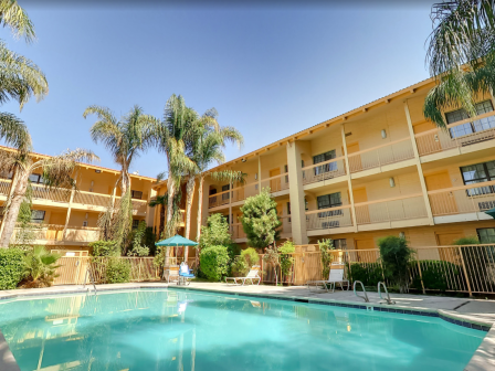 A swimming pool surrounded by palm trees and multi-story buildings with balconies, featuring umbrellas and lounge chairs in a sunny setting.