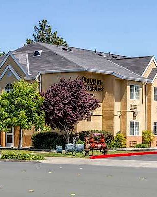 The image shows a yellow two-story hotel building with a sign that reads "Quality Inn & Suites" surrounded by trees and a clear sky.
